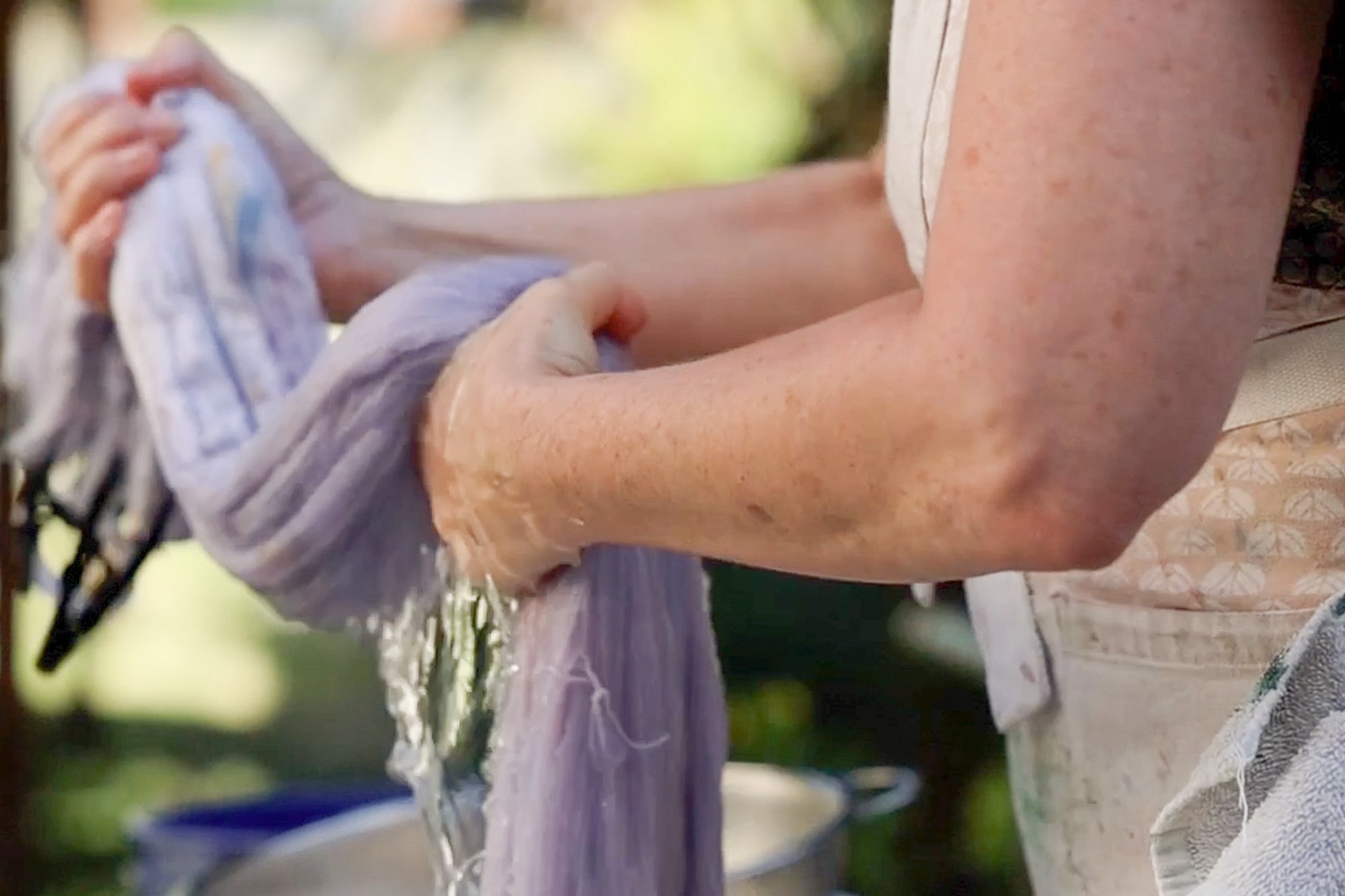 Anne, a white woman, is hand-washing purple yarn in a bucket. She's squeezing the wet yarn and drops of water are visible.