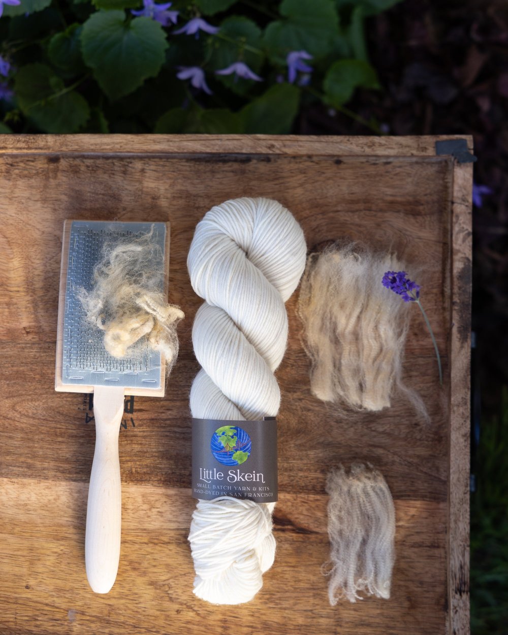 Flatlay showing carding brush with wool on it, wound skein of undyed yarn and representative samples of the fiber contained in the yarn: merino and rambouillet wool