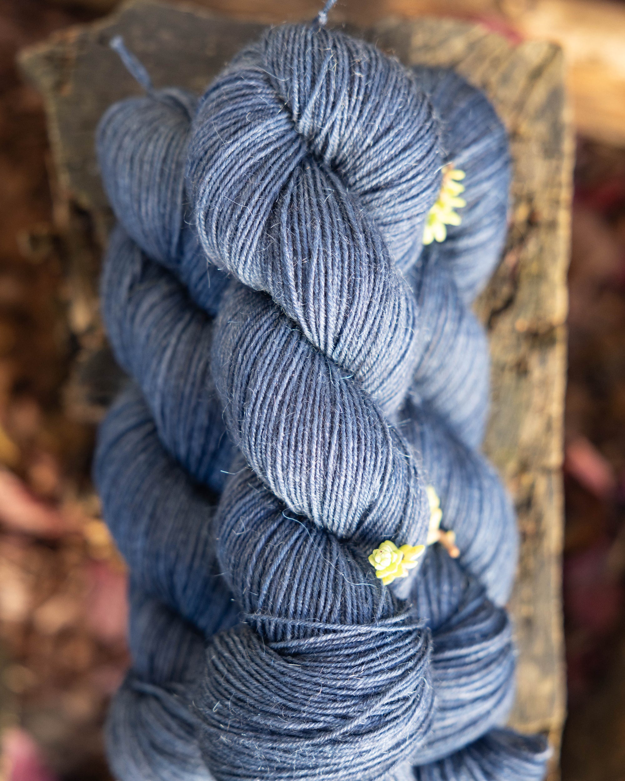 A small stack of deep blue colored hand-dyed yarn, placed on a wood stump and with pieces of succulents scattered around.